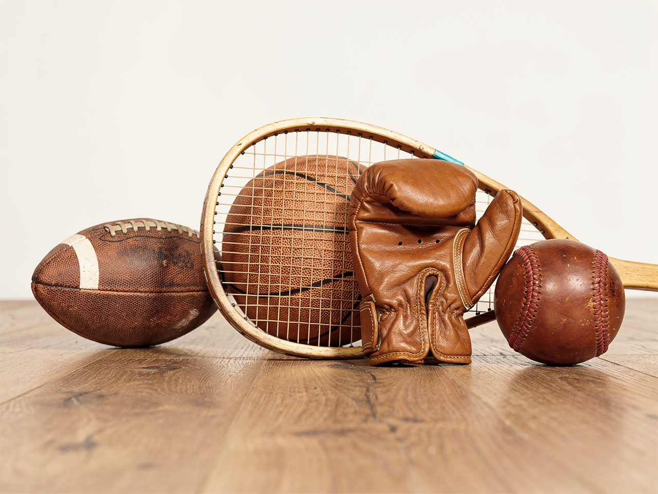 Vintage sports gear, including a football, basketball, wooden racket, boxing glove, and baseball, sits on a wooden floor.