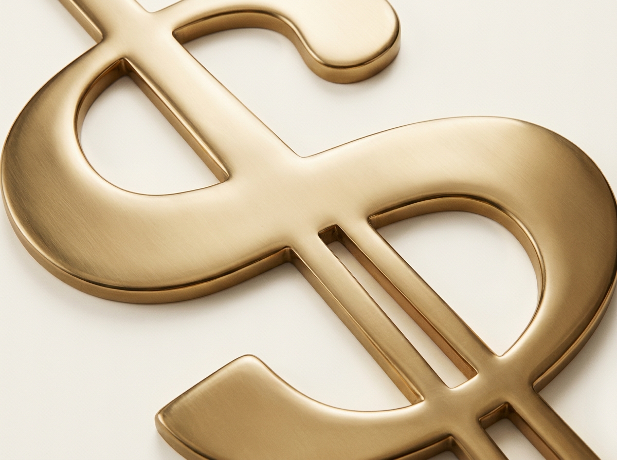A brushed brass dollar sign with double vertical bars lies on an off-white background in a close-up, angled view.