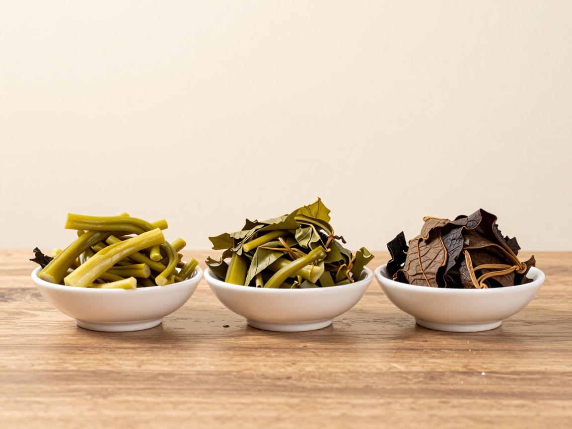 Three small white bowls on a wooden table hold pickled leaves and stems in pale yellow-green, olive green and dark brown.